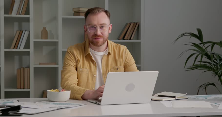 Man in stylish outfit working from office, using laptop, looking at camera. Bowl with colorful cereals on the desk near paper documents. Man with attractive appearance and nice smile. | Shutterstock HD Video #1111086823