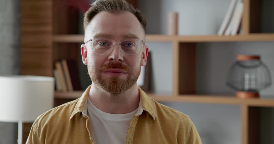 Smiling confident man with eyeglasses looking in camera, standing in front. Man in stylish outfit with ginger beard posing on camera. Modern interior design of room, shelves with stylish details | Shutterstock HD Video #1111086825