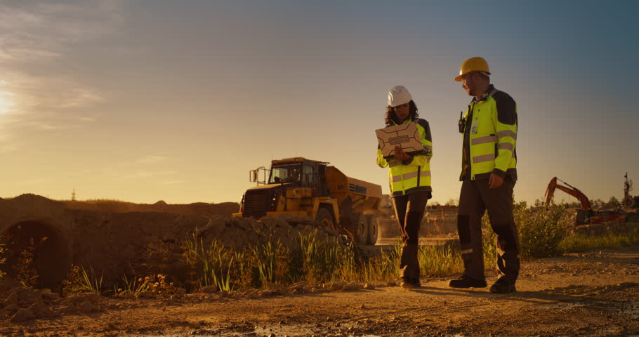 Caucasian Male Architect And Hispanic Female Urban Planner Walking On Construction Site With Laptop Computer And Talking About New Real Estate Project. Construction Truck Driving By. Golden Hour Royalty-Free Stock Footage #1111090481