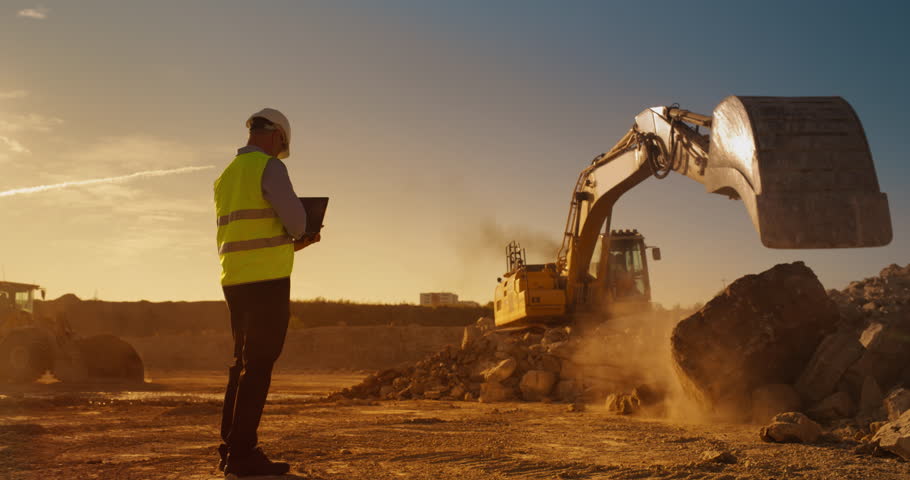 Caucasian Male Inspector Using Laptop Computer On Construction Site Of New Real Estate Project. Man Observing Industrial Machinery At Work, Laying Foundation For Apartment Building. Sunny Evening