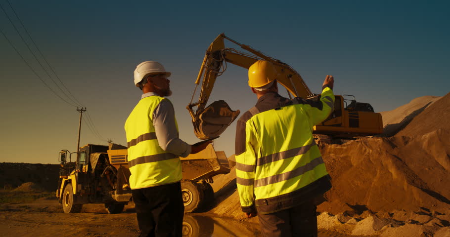 Zoom Out Back View Of Caucasian Real Estate Investor With Tablet Computer And Project Manager Talking On Construction Site. Colleagues Wearing Hard Hats And Watching Heavy Machinery Working. Sunny Day