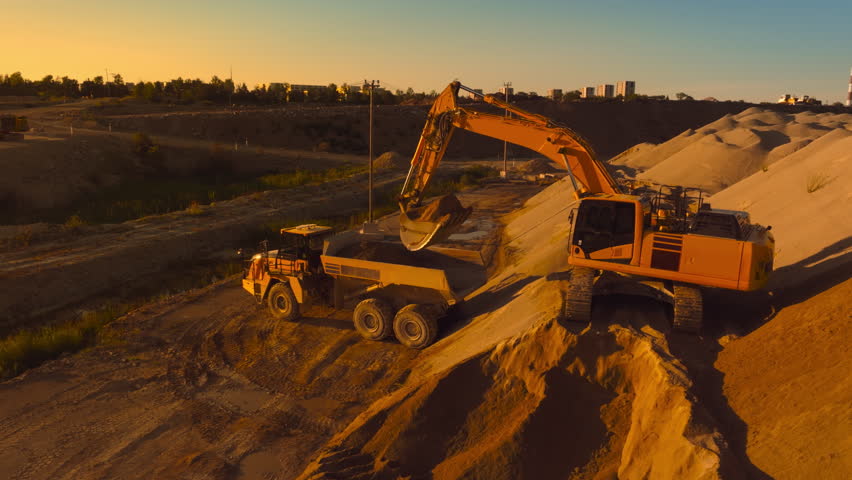 Aerial Drone Shot Of Construction Site On Sunny Day: Industrial Excavator Loading Sand Into A Truck. The Process Of Building New Apartment Complex. Workers Operating Heavy Machinery Concept.