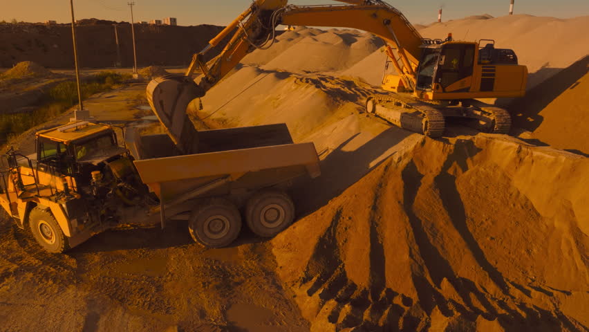 Aerial Drone Shot Of Construction Site On Sunny Evening: Industrial Excavator Loading Sand Into A Truck. The Process Of Building New Apartment Complex. Workers Operating Heavy Machinery Concept.