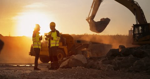 Cinematic Golden Hour Shot Of Construction Site: Caucasian Male Civil Engineer And Hispanic Female Architect Talking And Using Tablet. Trucks, Excavators, Loaders Working To Build New Apartment Block.: stockvideo