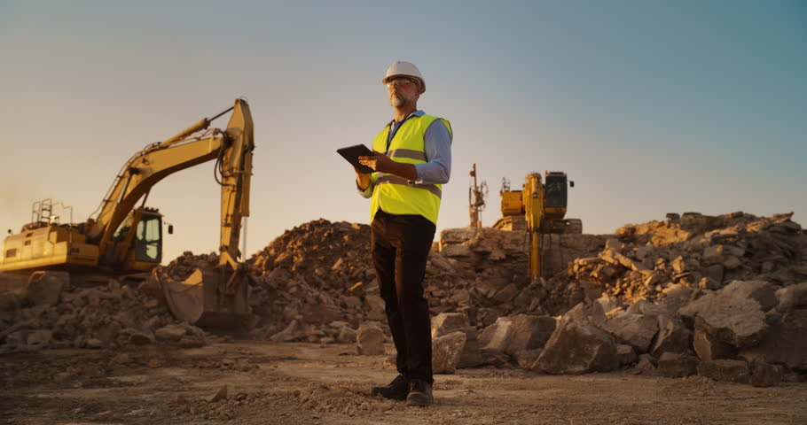 Caucasian Male Real Estate Investor Wearing Protective Goggles And Using Tablet On Construction Site On A Sunny Day. Man Inspecting Building Progress. Excavator Loading Materials Into Industrial Truck