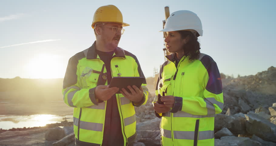 Caucasian Male Civil Engineer Talking To Hispanic Female Architect And Using Tablet On Construction Site of New Apartment Building. Real Estate Developers Discussing Business, Excavators Working.