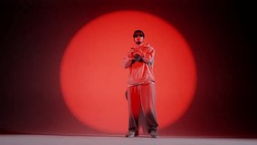 In the frame on a red background stands a young man on it shines a spotlight beam. Highlights him in a circle. Demonstrates a dance movement, jumping. Dressed in street style clothing. Slow motion