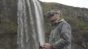Man controls an FPV drone for aerial photography and video with a remote control and goggles, behind a waterfall in Iceland