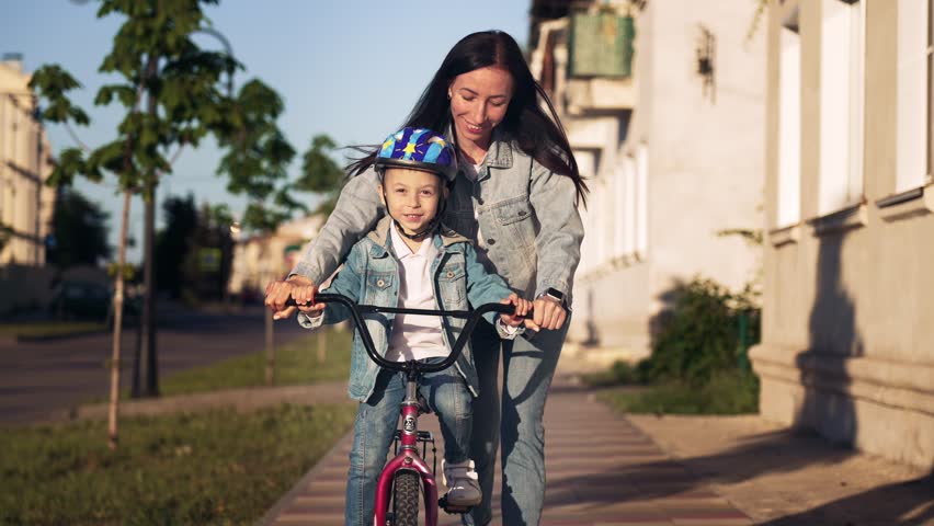 Happy family walk on street the city mother teaches her son child to ride a bike and run nearby. Slow motion woman helps keep balance for safe activity. Sports walk in nature. Healthy lifestyle. Royalty-Free Stock Footage #1111096319