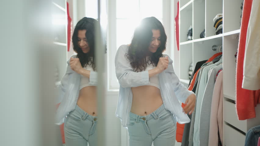 Beautiful smiling brunette in jeans and white top puts on light shirt in a walk-in closet at home. Young woman poses for camera. Feel like a fashion model. High quality 4k footage