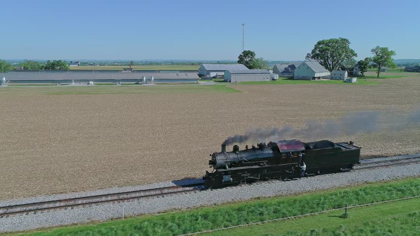 A drone view over a steaming train passing on a railroad between rural fields