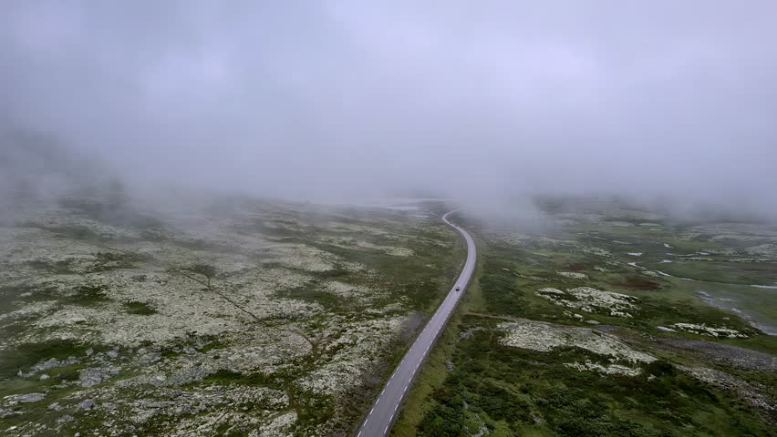 Aerial view flying over a car driving on a winding mountain road along mountain slope in the foggy weather in Norway. Rondane National Park in the Norwegian nature tundra landscape