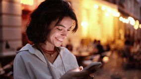 Smiling woman using smartphone on street with night city lights on background. Hipster female blogger typing text message on her cellphone - Powered by Shutterstock - Get 15% off with code: PIKWIZARD15