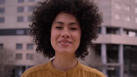 Young beautiful African American girl smiling looking at camera and afro hair. Cheerful and happy millennial woman posing for video. Latin people with positive expression outdoors.