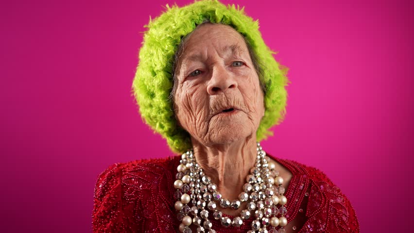 Funny, elderly mature woman, 80s, giving peace sign gesture with no teeth and green hat, isolated on pink background studio. Concept of youthful old lady Royalty-Free Stock Footage #1111108699