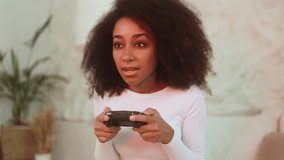 Portrait of excited curly woman gamer sitting on a bed and playing in video games on a console at home bedroom Happy player female controlling joystick for online competition indoors