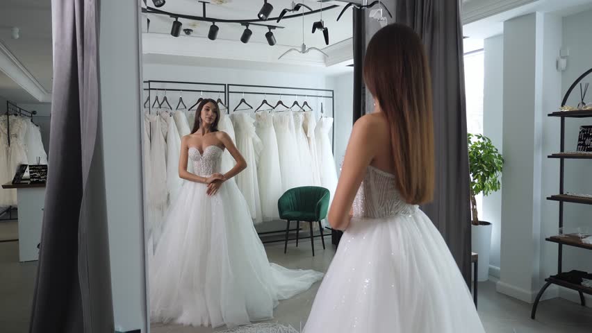 A beautiful bride chooses a wedding dress in a wedding salon, she stands in front of a mirror and turns from side to side. Royalty-Free Stock Footage #1111119557