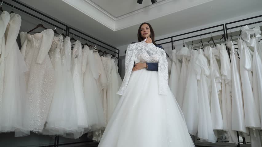 Portrait of a happy bride with long dark hair in a wedding salon, she holds a beautiful white wedding dress in her hands and dances. There are a lot of wedding dresses on hangers in the background. Royalty-Free Stock Footage #1111119559