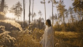 Outdoor slow motion video of young beautiful lady in autumn landscape with dry wild flowers and tall grass. light white dress. brunette woman walking in forest, park. warm autumn or summer season day