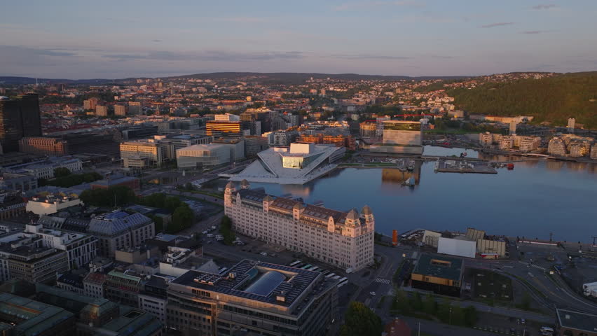 Aerial slide and pan footage of buildings in urban borough lit by setting sun. Modern design building of Operahuset Oslo on waterfront. Oslo, Norway