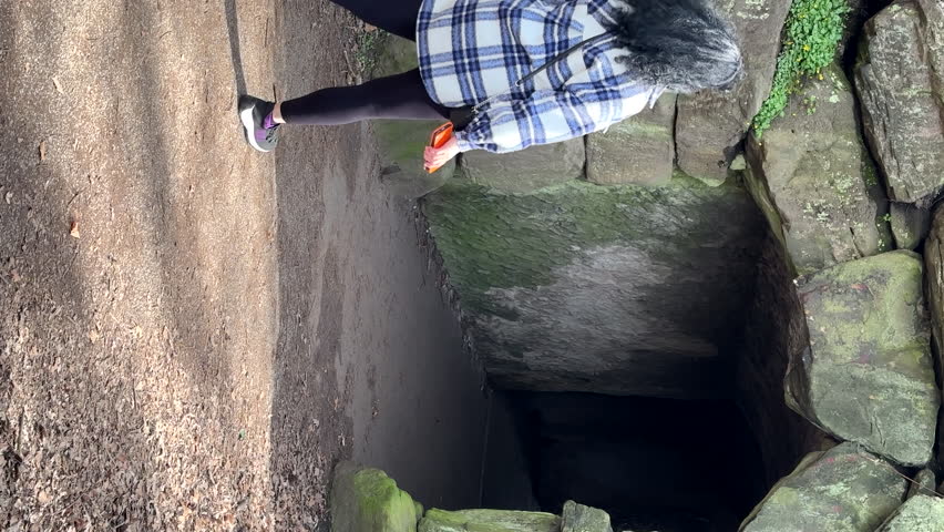 Vertical video. Rear view of long dark curly hair woman wearing checked pattern jacket and leggings entering and disappearing in arched stone tunnel.