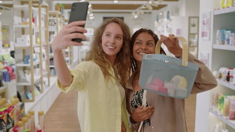 Medium long shot of two diverse female friends taking selfie with shopping basket while buying products at cosmetics store Vídeo Stock