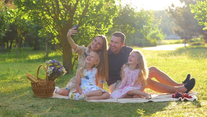 Happy family having picnic sitting on blanket in park on sunny day and taking selfie photos on smartphone. Young cheerful parents taking pictures with kids on mobile phone outdoor. Real time. Royalty-Free Stock Footage #1111140535