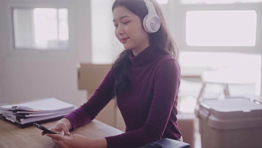young Asian student, dressed casually, enjoys listening to music through her headphones, selecting songs from her mobile phone and humming along at her desk in the dormitory Royalty-Free Stock Footage #1111144557