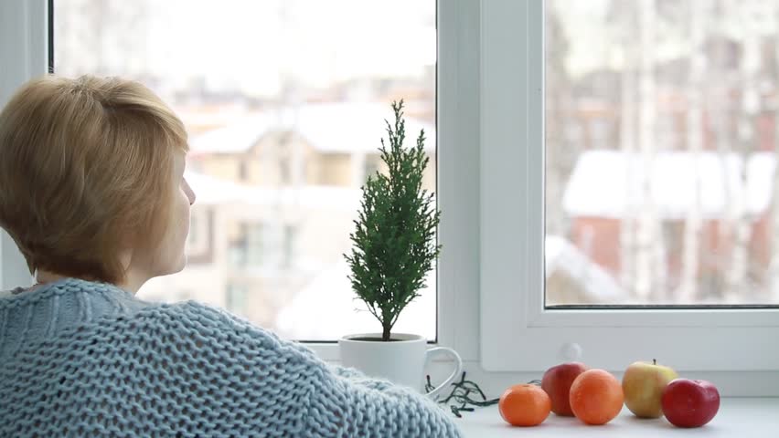 A woman sitting by the window next to fruit and a small Christmas tree growing in a cup on the windowsill
