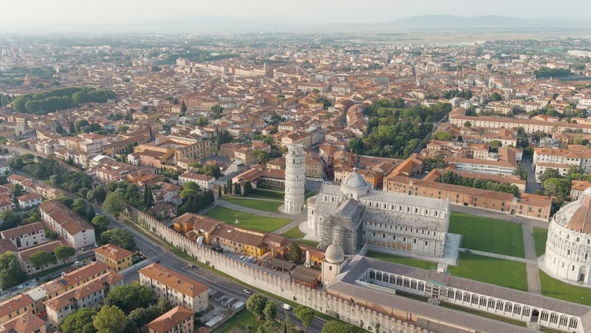 Pisa, Italy. The Leaning Tower of Pisa rises beside the Pisa Cathedral within the Piazza dei Miracoli, a UNESCO World Heritage Site. The cylindrical white marble bell tower, built in the Romanesque style with tiered arcades and loggias, leans at approximately 4 degrees due to the soft subsoil beneath its foundation. The cathedral and baptistery stand nearby on manicured green lawns, surrounded by the historic city center with terracotta rooftops. The scene is captured in warm morning light during summer, with clear skies and soft atmospheric haze over the distant Tuscan landscape. Aerial view, Point of interest