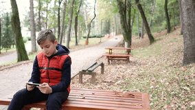 Cute schoolboy talking with his friends on mobile phone. Boy is sitting on the bench in public park in autumn.