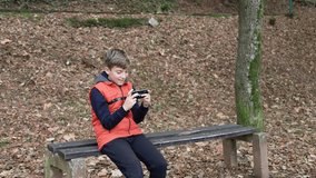 Cute schoolboy playing video games on his mobile phone. Boy is sitting on the bench in public park in autumn. He is unhappy because he is loosing.