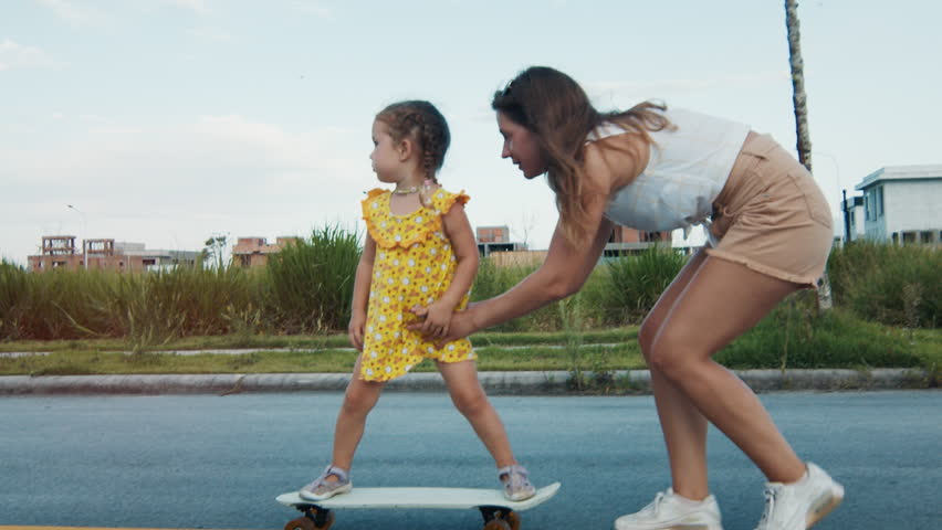 Woman teaches little girl how to skateboard on the asphalt road. Happy girl in summer dress enjoys riding skate on the street