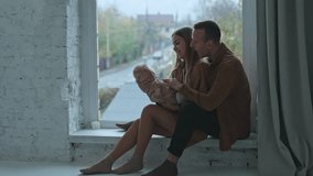 Young couple sitting on the window-sill with baby in hands. Loving parents talk to the child and calm him down. - Powered by Shutterstock - Get 15% off with code: PIKWIZARD15