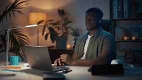 Positive african american man in wireless earphones sitting at home office and using laptop for video call. Male freelancer leaning on comfy chair and relaxing after remote work at evening time.