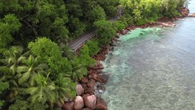 Bird eye drone shot of passing bus on asphalt road, granite rocks, trees and turquoise water on the baie lazare shore, Mahe Seychelles 30 fps
