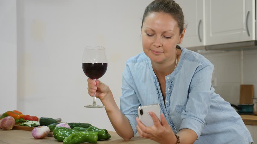 Adult European female holidng a glass of wine and a phone at the kitchen counter at home