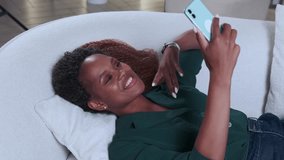 Young cheerful pretty African American woman using phone for video chatting and enjoying opportunity to relax and chat with friends after hard day of work lies on white sofa at home.