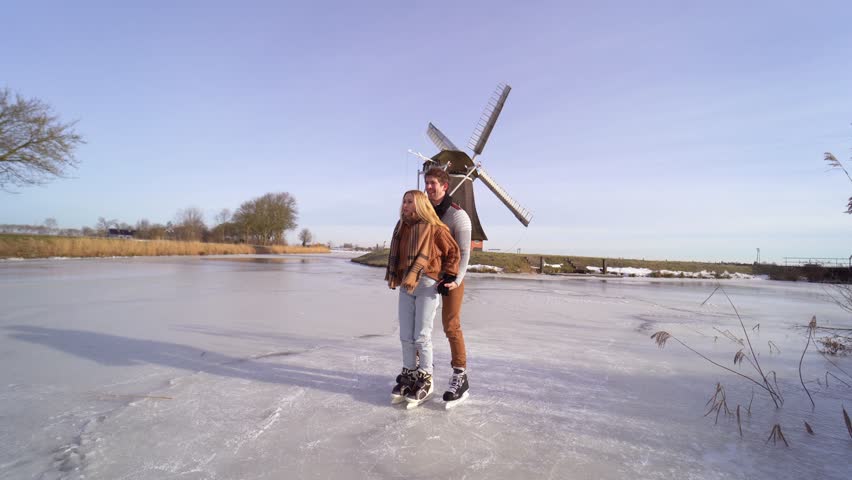 Loving couple having fun on ice in typical dutch landscape with windmill. Woman and man ice skating outdoors in sunny snowy day. Romantic Active date on frozen canal in winter Christmas Eve. | Shutterstock HD Video #1111187415