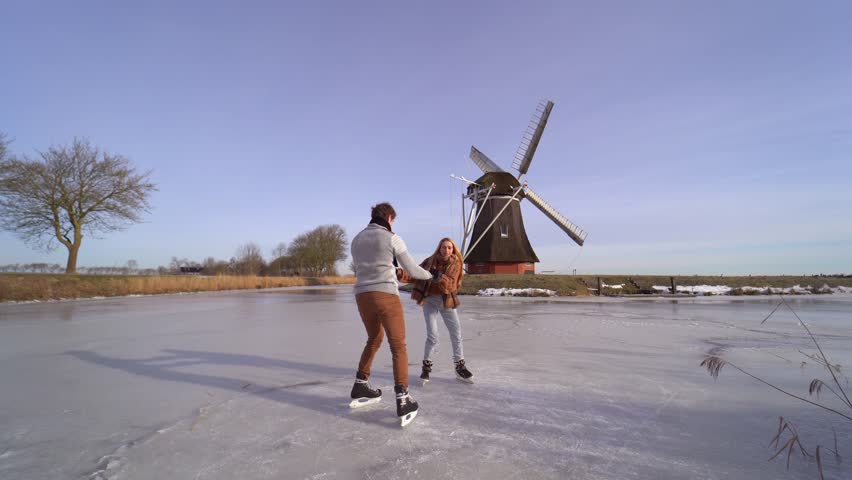Loving couple having fun on ice in typical dutch landscape with windmill. Woman and man ice skating outdoors in sunny snowy day. Romantic Active date on frozen canal in winter Christmas Eve. | Shutterstock HD Video #1111187417