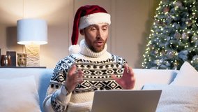 Handsome Caucasian man with Santa Claus hat talking on video call on his laptop in modern living room with Christmas tree in background. Remotely congratulating his family or friends on holidays.