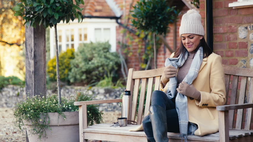 Portrait of woman dressed for autumn or winter walk outside house sitting on bench with flask of hot drink - shot in slow motion