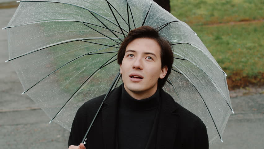 Young man in a coat putting away the umbrella, exposing his face to the rain and holding his arms to the sides with a smile on his face in the autumn city park in rainy weather close up