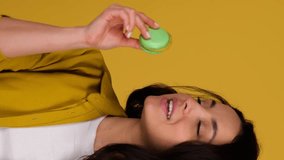 Happy pretty woman choosing between two macarons and biting one, posing in studio, isolated on yellow background. Sweets food concept. Real time vertical video