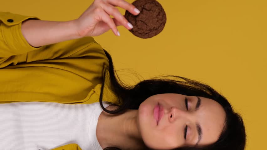 Happy young lady enjoying oat cookies while biting. Lady standing against yellow background. Healthy, junk food concept. Real time vertical video Royalty-Free Stock Footage #1111200689