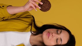 Happy young lady enjoying oat cookies while biting. Lady standing against yellow background. Healthy, junk food concept. Real time vertical video
