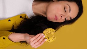 Smiling pretty lady choosing between two donuts and biting one, posing in studio, isolated on yellow background. Real time vertical video