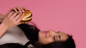 Happy woman biting a hamburger while standing and posing in studio over pink background. People, lifestyle, junk food concept. Real time vertical video