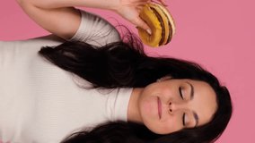 Happy young lady holding burger and dancing while posing in studio over pink background. People, lifestyle, junk food concept. Real time vertical video