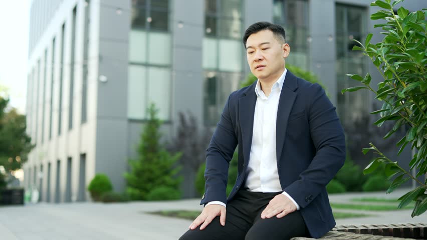 Tired overworked asian businessman in formal suit suffering from back pain sitting on a bench on the street near an office building. Upset exhausted entrepreneur massages painful lower back muscles Royalty-Free Stock Footage #1111210199
